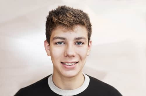 headshot of boy in dark t shirt smiling showing metal braces with solid grey background