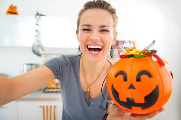 Mom in a kitchen decorated for Halloween smiling and holding up a plastic pumpkin full of candy